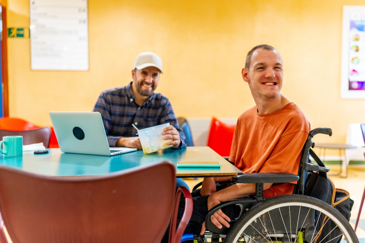 Man,With,Disability,And,Coworker,Eating,Together,In,The,Dinning man in wheelchair having lunch with coworker